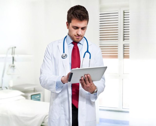 Male physician in exam room entering patient data on a medical tablet.