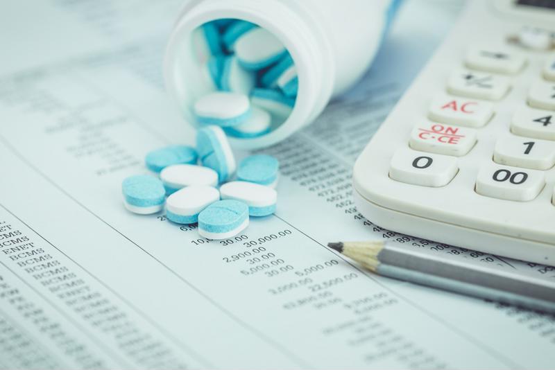 An open prescription bottle with blue and white pills spilling onto a spreadsheet next to a pencil and calculator.